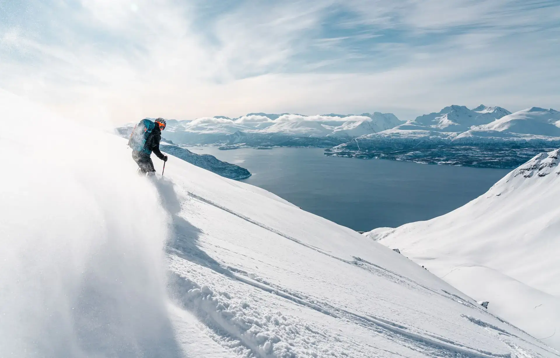 A skier descending a snowy slope above a fjord and mountain ridges